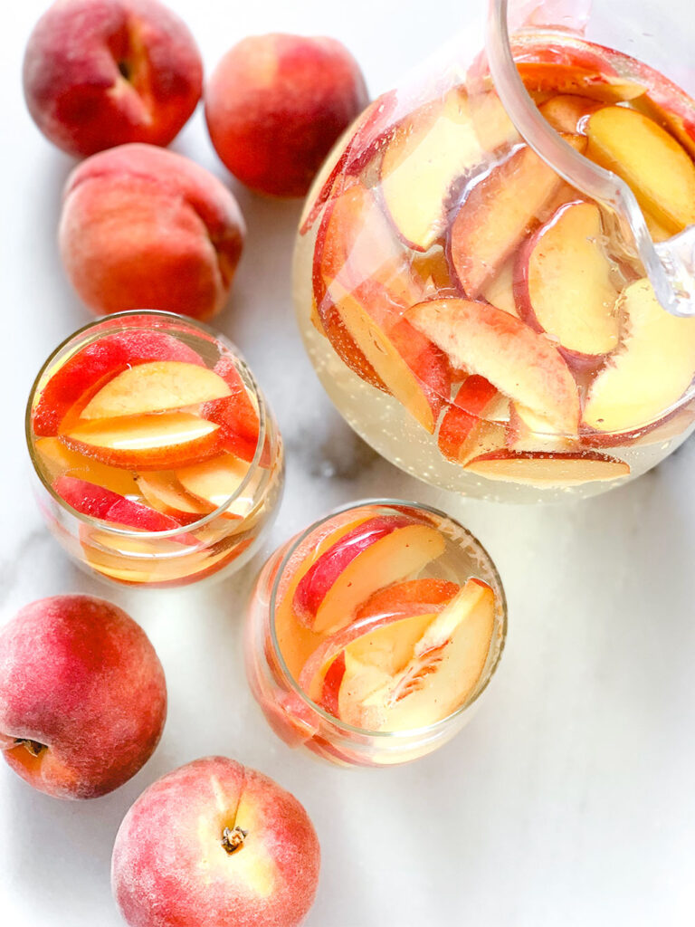 Peach Sangria in a pitcher and in stemless wine glasses with whole peaches on a table