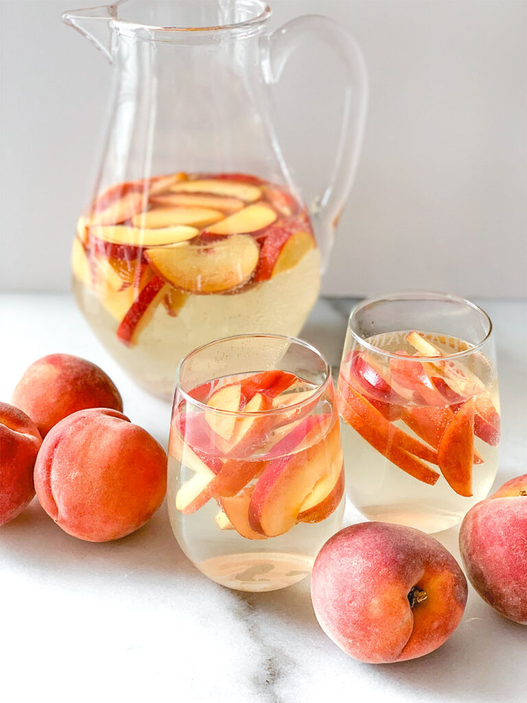 Peach Sangria in a pitcher and in stemless wine glasses with whole peaches on a table