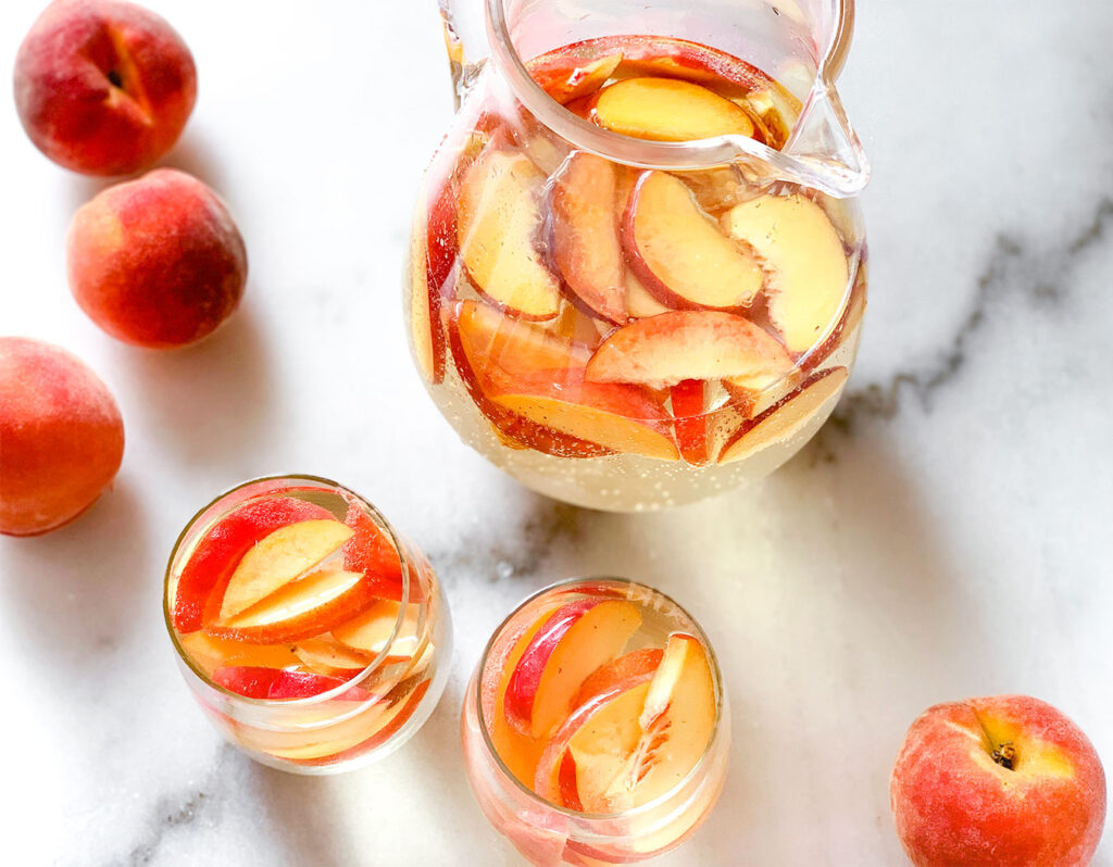 Peach Sangria in a pitcher and in stemless wine glasses with whole peaches on a table