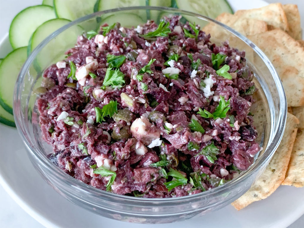 Kalamata Olive Tapenade in a bowl with sliced cucumbers and crackers