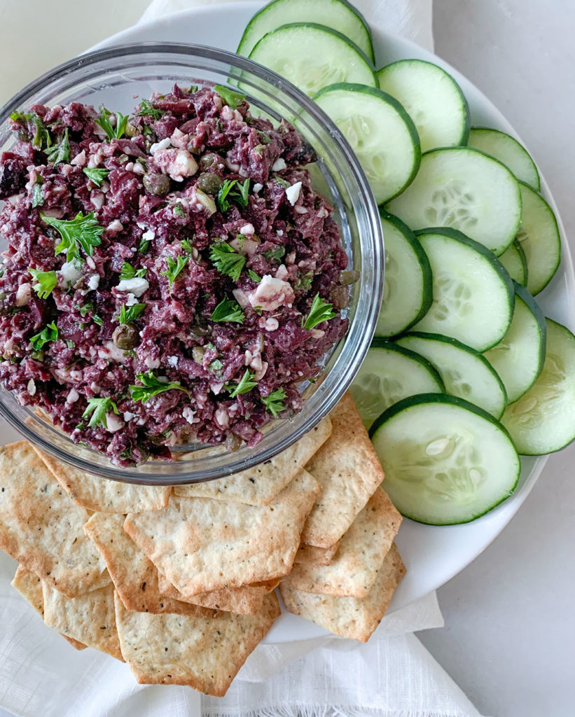 Kalamata Olive Tapenade in a bowl with sliced cucumbers and crackers