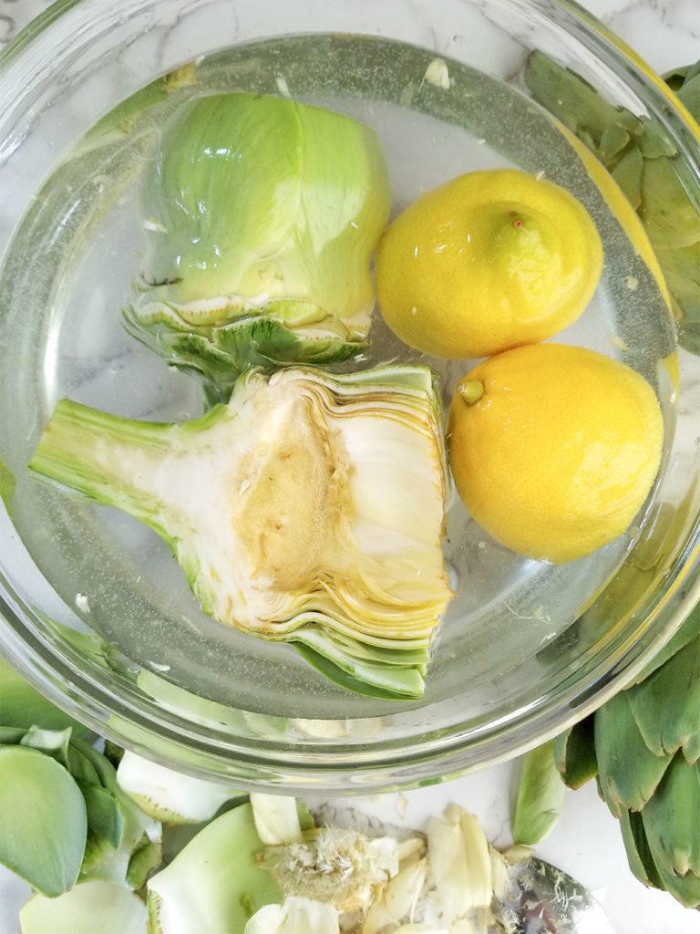 prepared artichokes soaking in a bowl of water with lemon