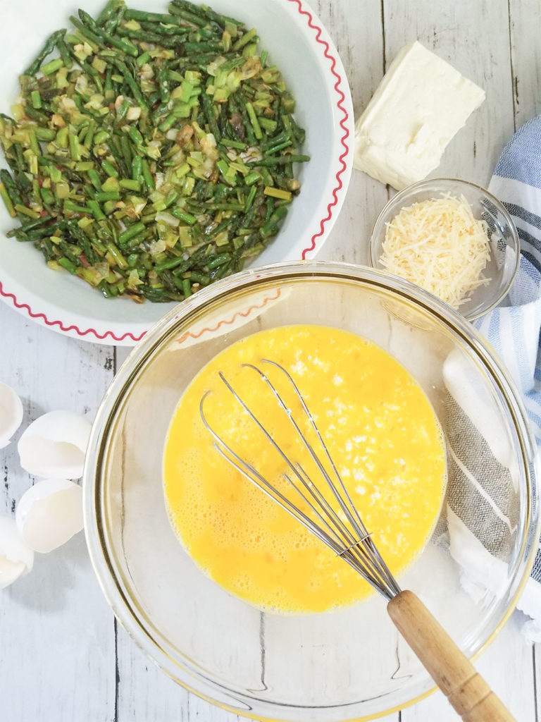 Sautéed asparagus and green onion in a pan, egg mixture in a bowl with a whisk