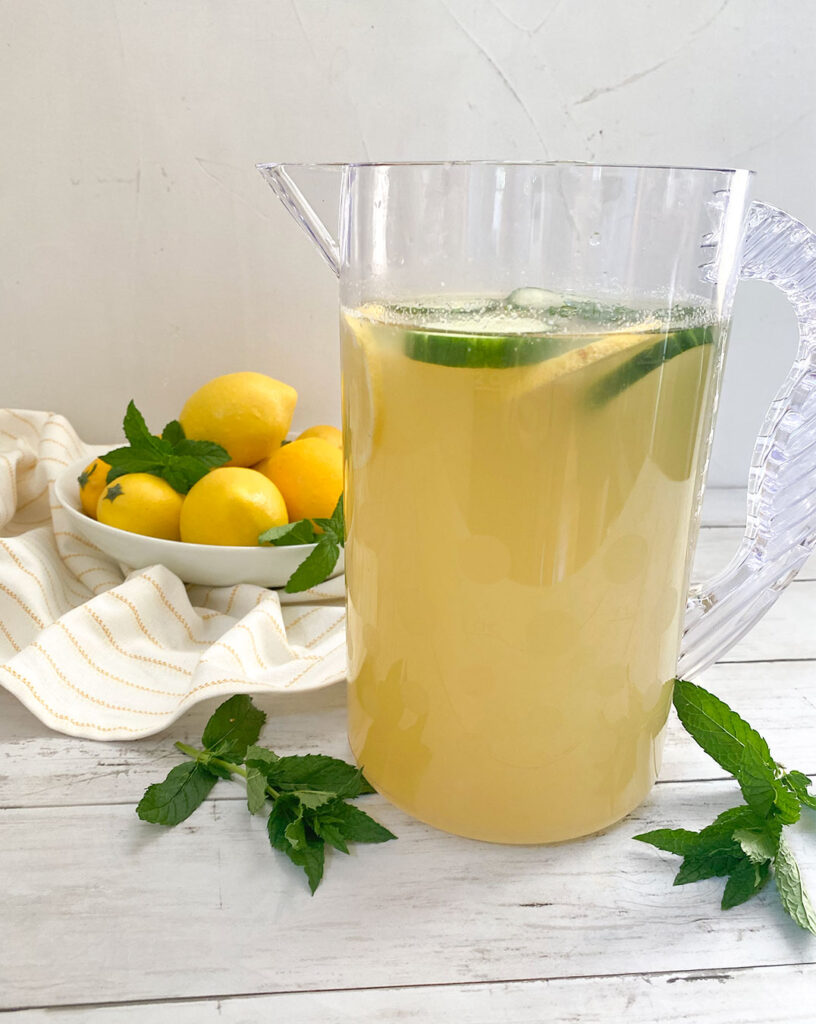 Cucumber Mint Lemonade in a pitcher with a bowl a lemons and mint sprigs