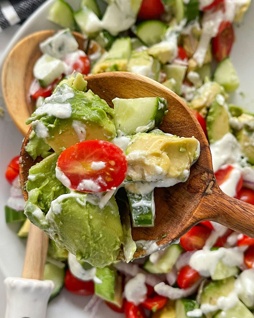 A wooden spoon with a scoop of Avocado, Cucumber, Tomato Salad with a creamy feta dressing. 
