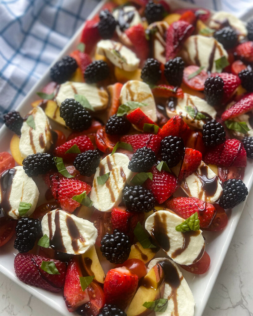 A white platter with a touch of a blue fabric around it,  With a Summer Fruit Caprese Salad on top.