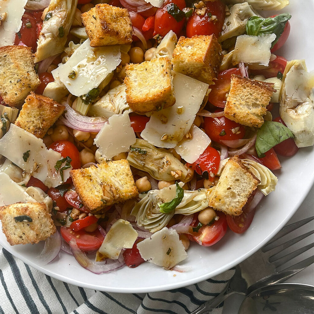 A white bowl filled with a delicious salad of marinated artichokes, sweet roasted red peppers, chickpeas, sliced red onion and topped with homemade crunchy croutons.