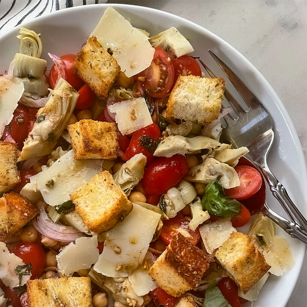 A delicious and colorful Mediterranean Artichoke Salad in a white ceramic bowl, featuring artichoke hearts, juicy cherry tomatoes, red onion slices, chick peas and homemade croutons all topped with shaved parmesan cheese, drizzled with vinaigrette dressing and garnished with chopped basil.