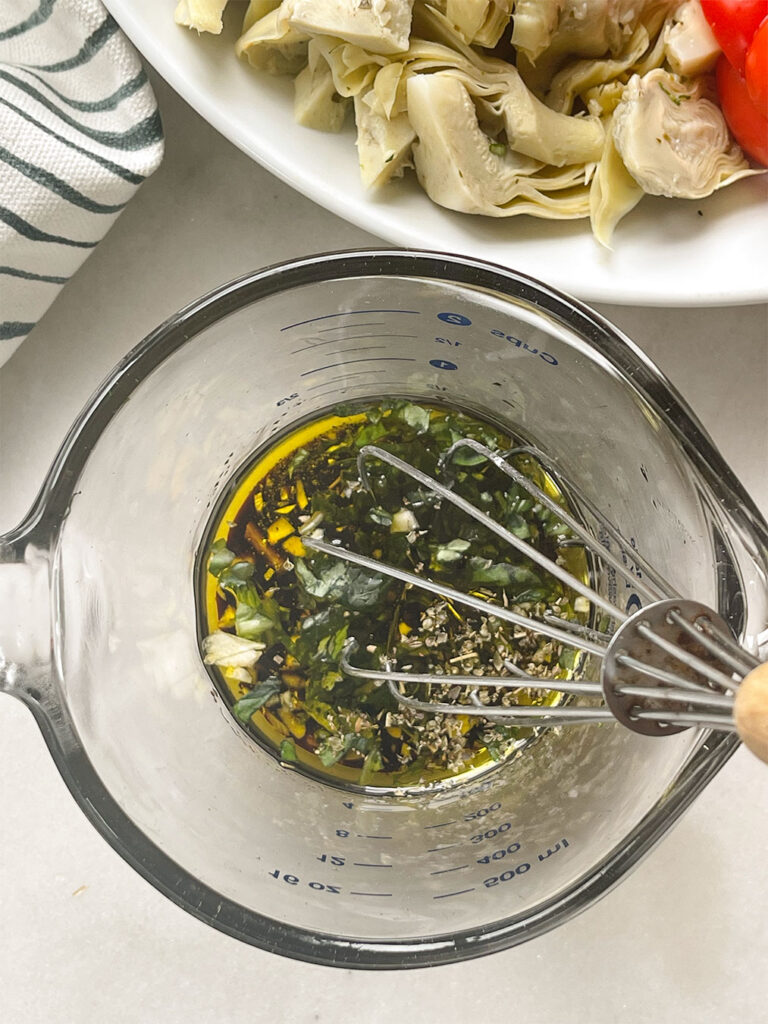 A glass measuring cup filled with a homemade dressing for a salad, made of high-quality olive oil, tangy balsamic vinegar, sweet honey, oregano, chopped garlic and freshly chopped basil, whisked together to form a flavorful and aromatic blend