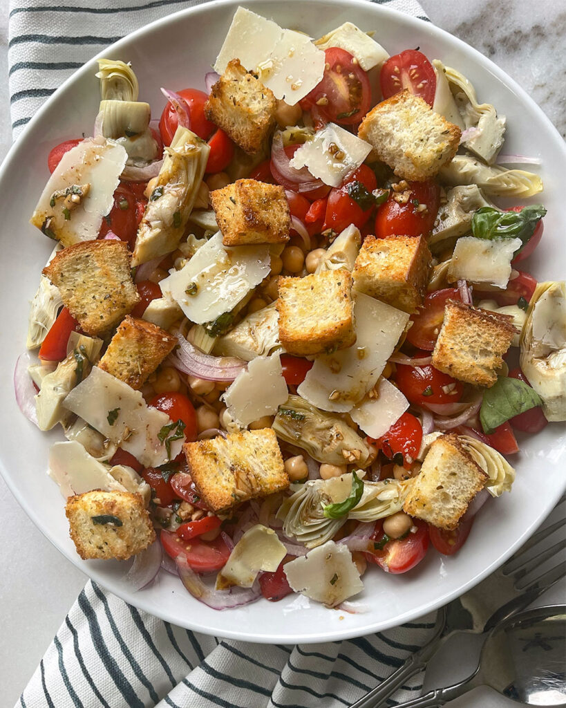 A bowl of Mediterranean Artichoke Salad filled with marinated artichokes, juicy cherry tomatoes, chickpeas, sweet re onion, roasted red peppers, all drizzled with a sweet vinaigrette and topped with shaved parmesan cheese.