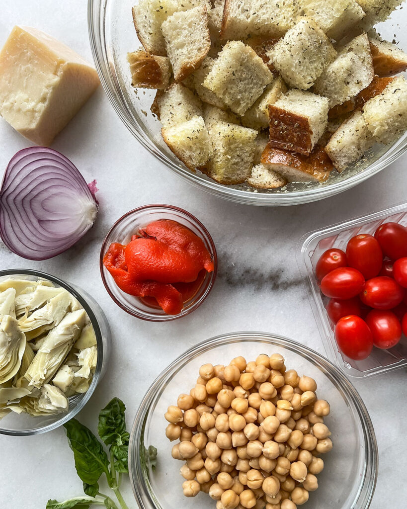 A arrangement of the ingredients to use in a Mediterranean Artichoke Salad, marinated artichokes, chickpeas, red onion, cherry tomatoes, and fresh basil.
