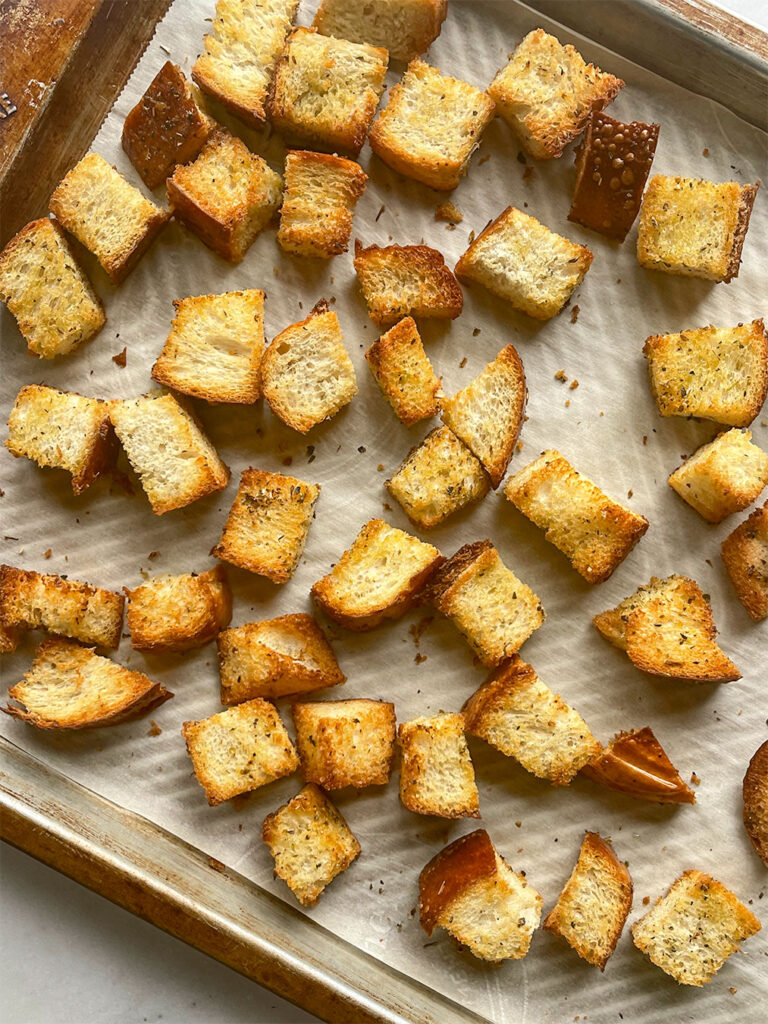 A baking pan filled woth cubes of Italian bread mixed in oilve oil and oregano ready to be baked. A baking sheet of the croutons freshly baked out of the oven.