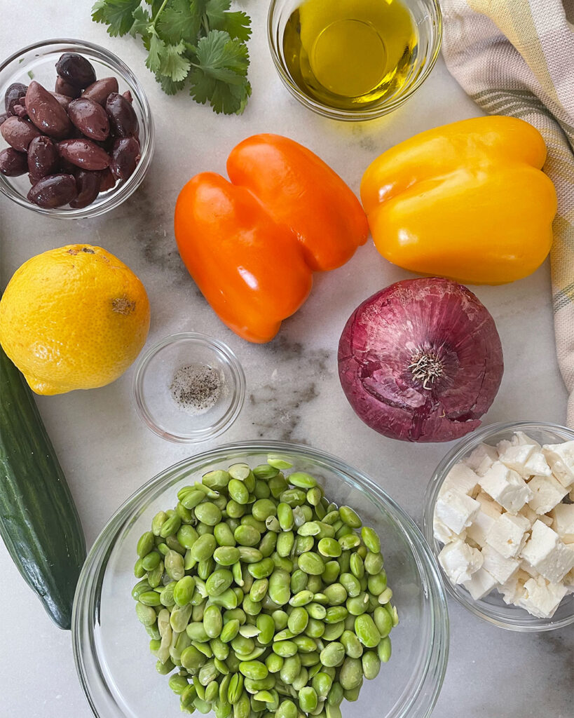 A colorful and appetizing array of ingredients for Mediterranean Edamame Salad, featuring  fresh cucumber, orange and yellow peppers, red onion, lemons, olive oil, feta cheese, and edamame beans .