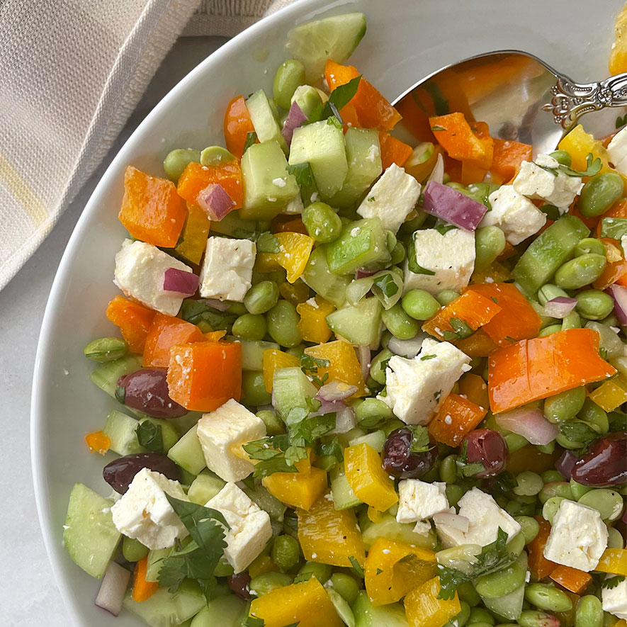 Close-up of a vibrant Mediterranean Edamame Salad featuring a mix of  colorful bell peppers, chopped cucumber, red onion slices, crumbled feta cheese, and Kalamata olives, arranged beautifully in a white bowl