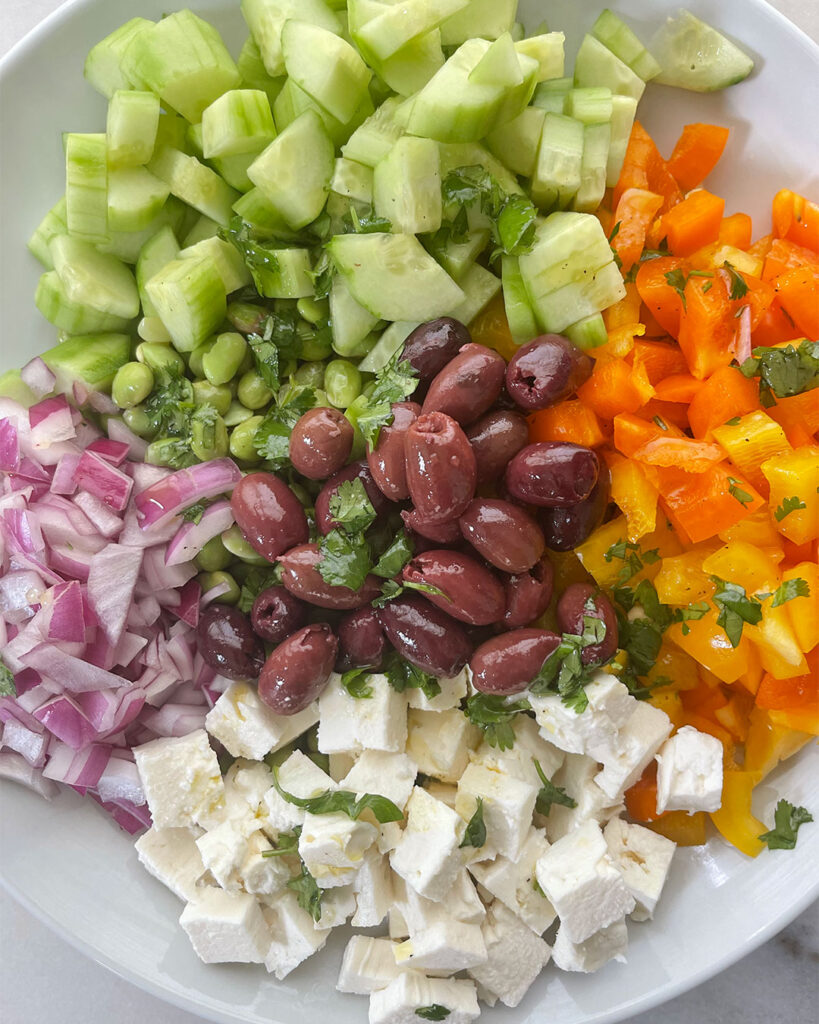 A bowl of green edamame followed by chopped cucumber, peppers  red onion. and cubed feta cheese.  Add Kalamtata olives to the top. 