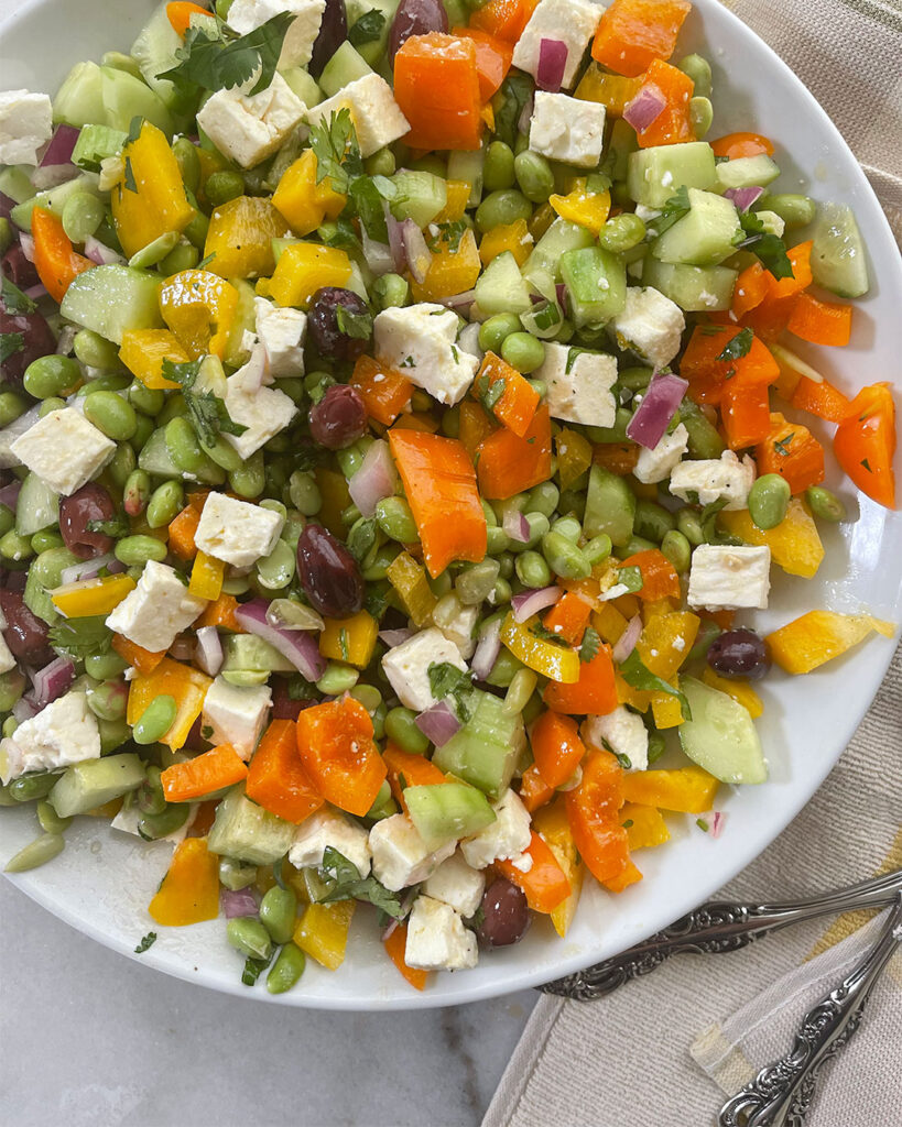 Close up of a colorful Edamame Salad featuring edamame, chopped peppers, cucumbers, red onion,  and Kalamata olives topped with feta cheese and  lemony Greek vinaigrette. 