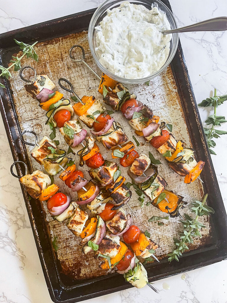 An appetizing tray of grilled Halloumi skewers, showcasing colorful peppers, zucchini ribbons, cherry tomatoes, and perfectly charred chunks of Halloumi cheese, ready to be enjoyed with a side of creamy tzatziki.