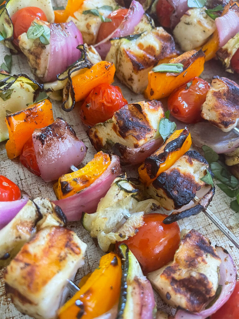 A mouthwatering close-up of Grilled Halloumi Skewers, featuring golden-brown charred edges of Halloumi cheese, vibrant colors of peppers, zucchini ribbons, and juicy cherry tomatoes, creating a tempting and irresistible grilled delight.