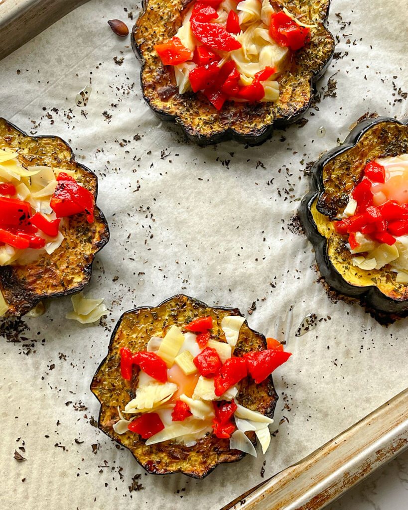 Baked acorn squash with an egg in the middle and artichokes and roasted red peppers on top on a sheet pan.