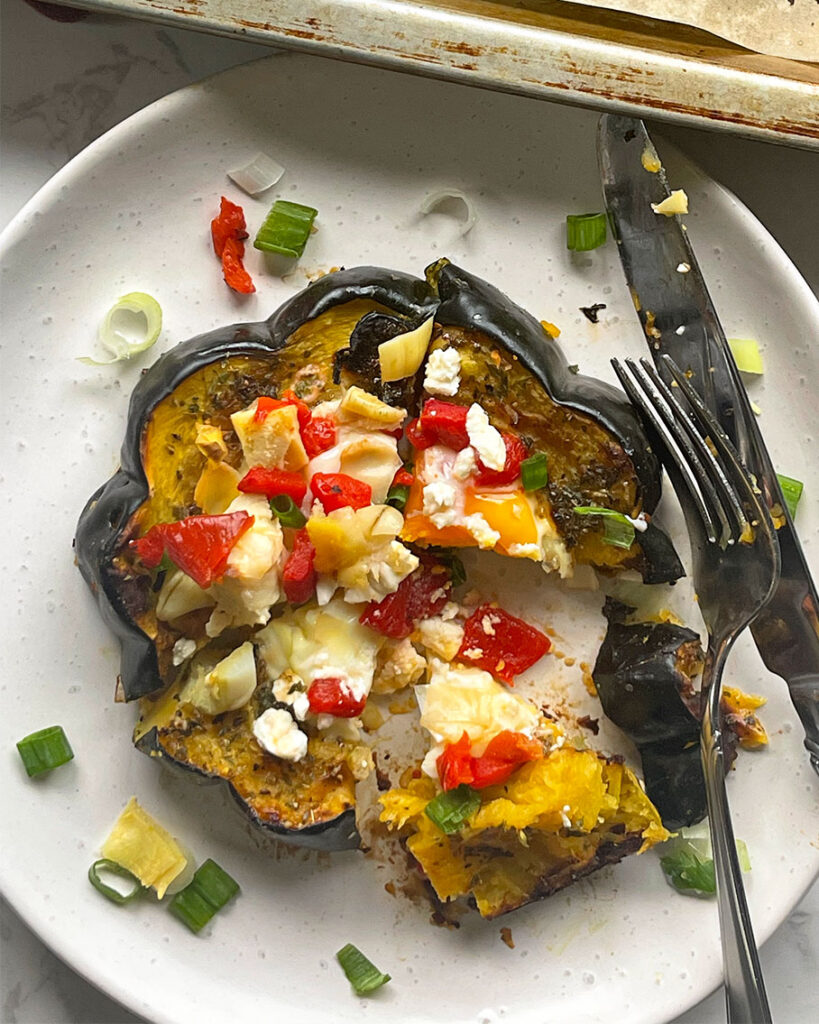 A white dish with a slice of acorn squash with a egg in the middle, topped with roasted red peppers, feta cheese, artichoke hearts and green onions.