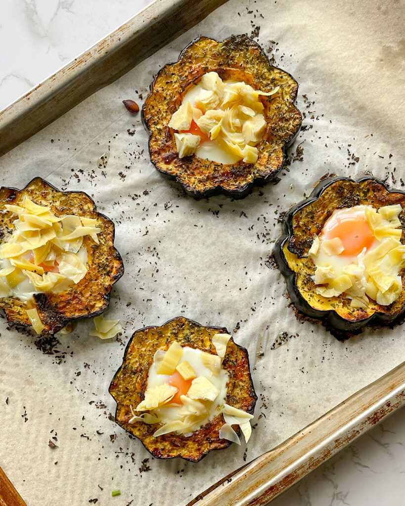 Baked acorn squash with an egg in the middle and artichoke on top on a sheet pan.