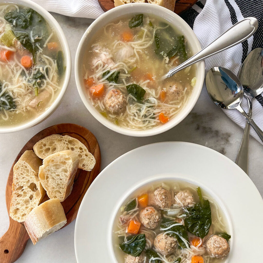 Steaming bowl of Italian Wedding Soup, a comforting mix of savory meatballs, tender vegetables, and hearty broth, served in a pristine white bowl alongside slices of crusty bread