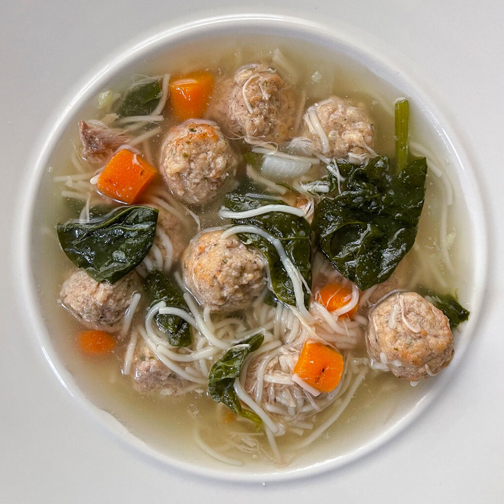 Steaming bowl of Italian Wedding Soup, a comforting mix of savory meatballs, tender vegetables, and hearty broth, served in a pristine white bowl.