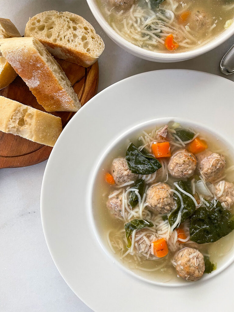 Steaming bowl of Italian Wedding Soup, a comforting mix of savory meatballs, tender vegetables, and hearty broth, served in a pristine white bowl alongside slices of crusty bread