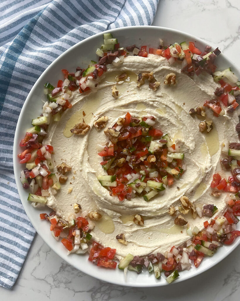 A bowl of tantalizing Gigantes Dip topped with vibrant grape tomatoes, crisp cucumber, and flavorful Kalamata olives, promising a burst of Mediterranean freshness in every bite