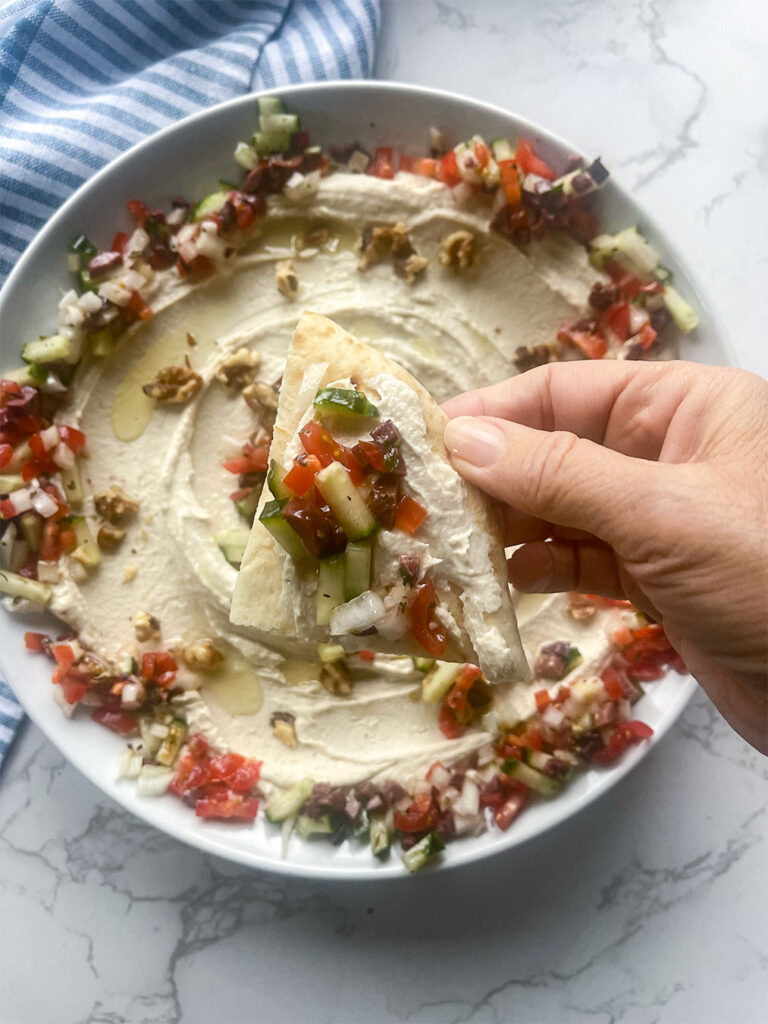 A hand dipping a piece of pita bread into a luscious bowl of Gigantes Dip, showcasing the creamy texture and enticing Mediterranean flavors.